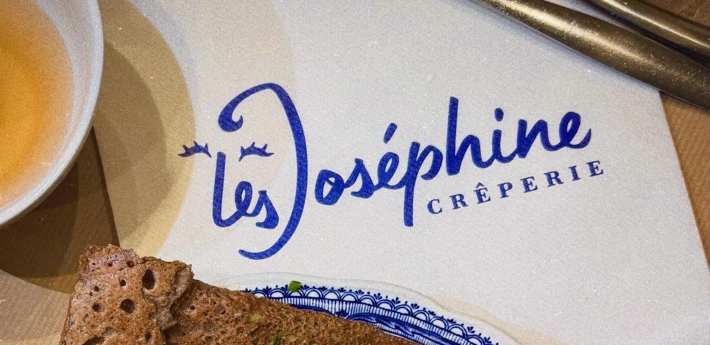 Logo bleu de la Crêperie Les Joséphine sur une serviette, avec une galette bretonne et une bolée de cidre en arrière-plan.